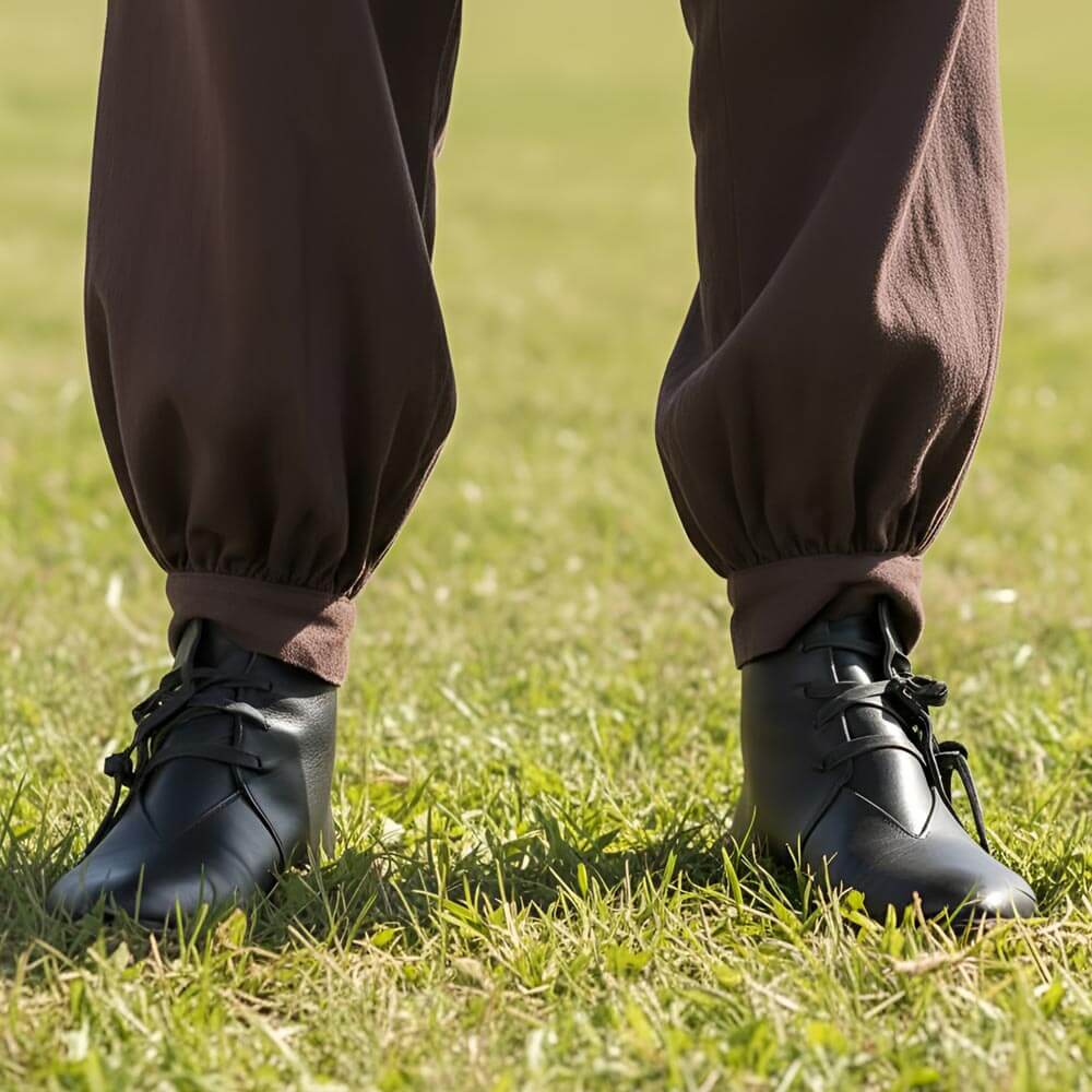 Black leather Renaissance ankle boots with four-string lacing, worn with brown pants on grass.
