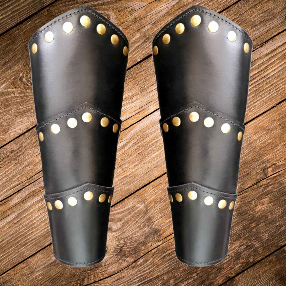 Pair of black leather Renaissance vambraces with gold rivet detailing on a wooden background.