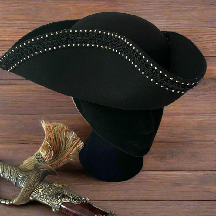 Black Renaissance pirate tricorn hat with gold trim and studs, displayed on a stand next to a sword.