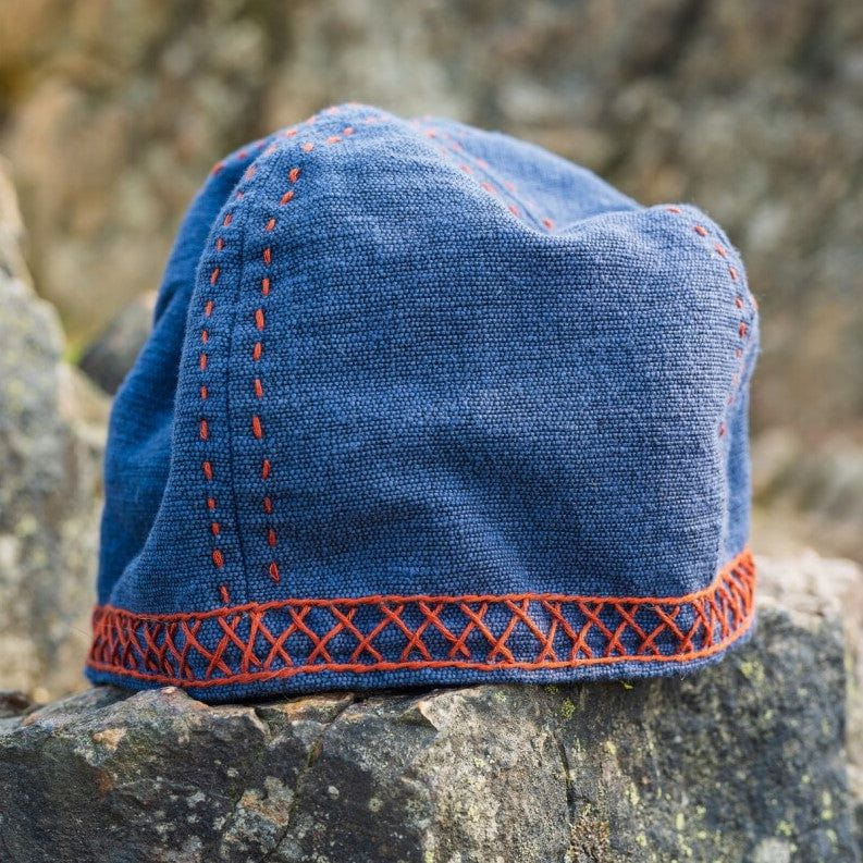 Blue cotton Renaissance cap with red embroidered geometric stitching on a rocky surface.