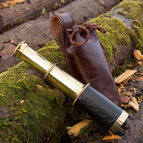 Renaissance brass spyglass with black leather grip and brown leather case on mossy logs.
