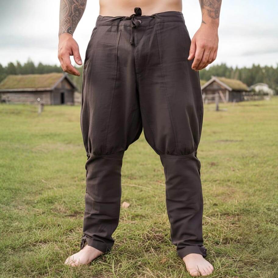 Person wearing brown Renaissance pants standing in a grassy field with wooden buildings in the background.