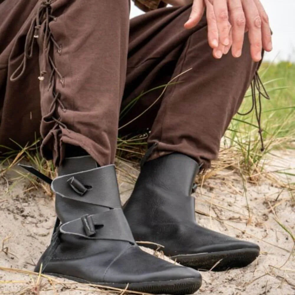 Black Renaissance leather ankle boots with toggle closures, worn with brown pants on sand.