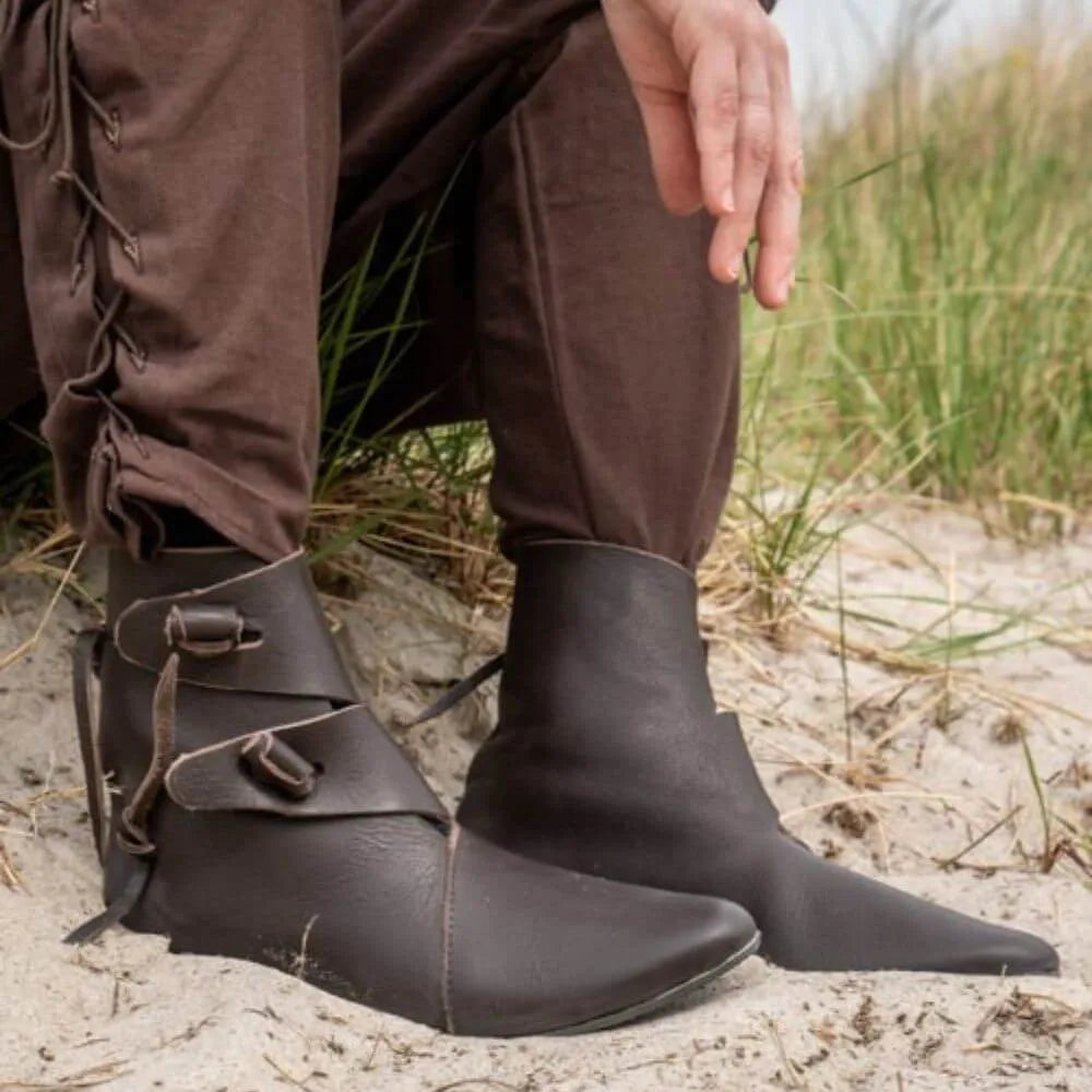 Brown Jorik Renaissance leather ankle boots with toggle closures and brown pants on sand.