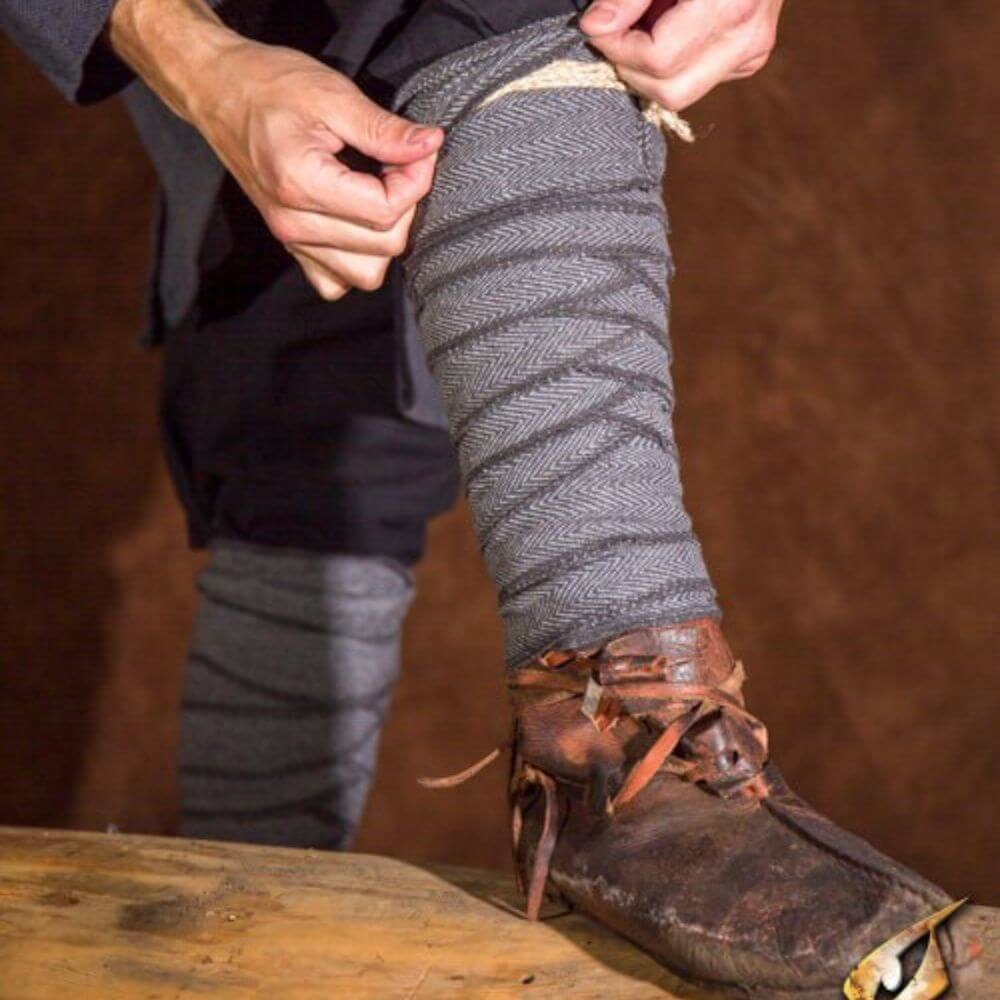 Man adjusts grey herringbone Leg Wraps - Protective Period Calf Wraps over trousers, wearing worn leather shoes.