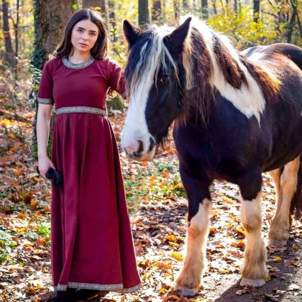 A woman dressed in a red short sleeve Viking dress stands in golden autumn woods with a draft horse.
