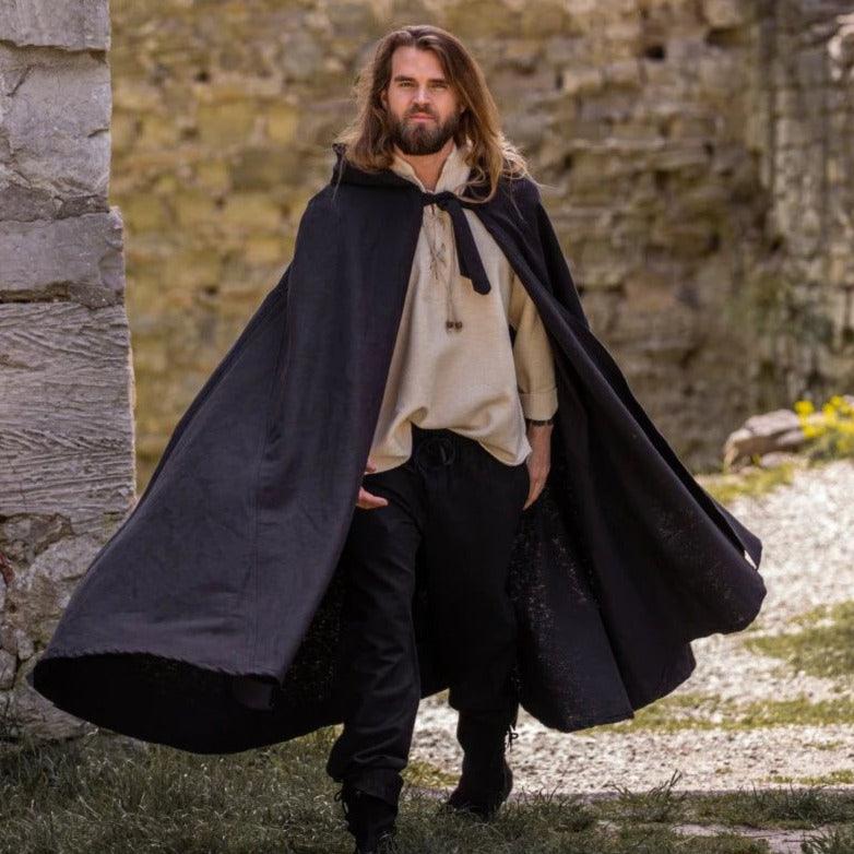 Man in a full-length black Renaissance woolen cloak with hood and front tie closure, walking outdoors.