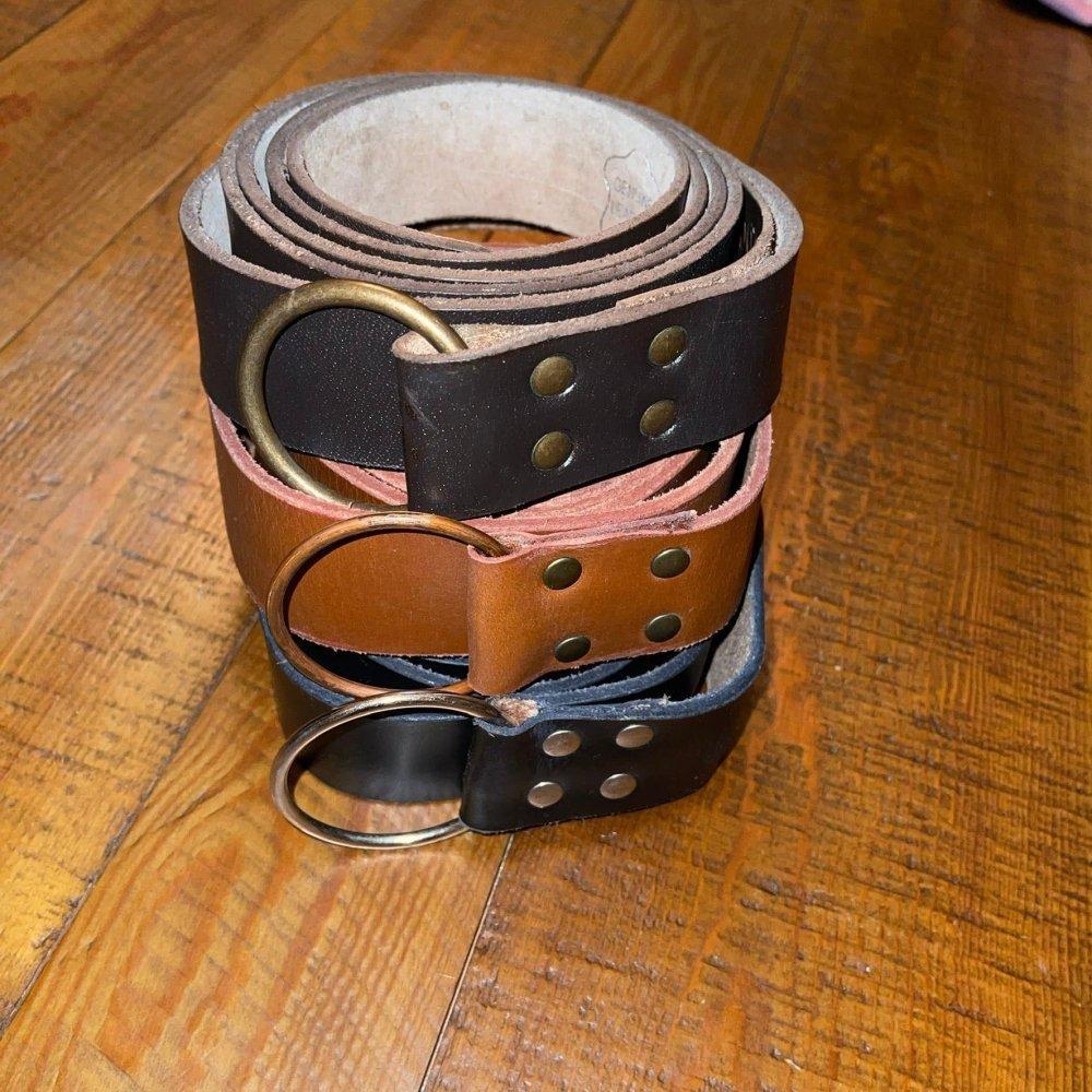 Stacked full-grain leather Renaissance belts in dark brown, light brown, and black, each with a brass ring loop.