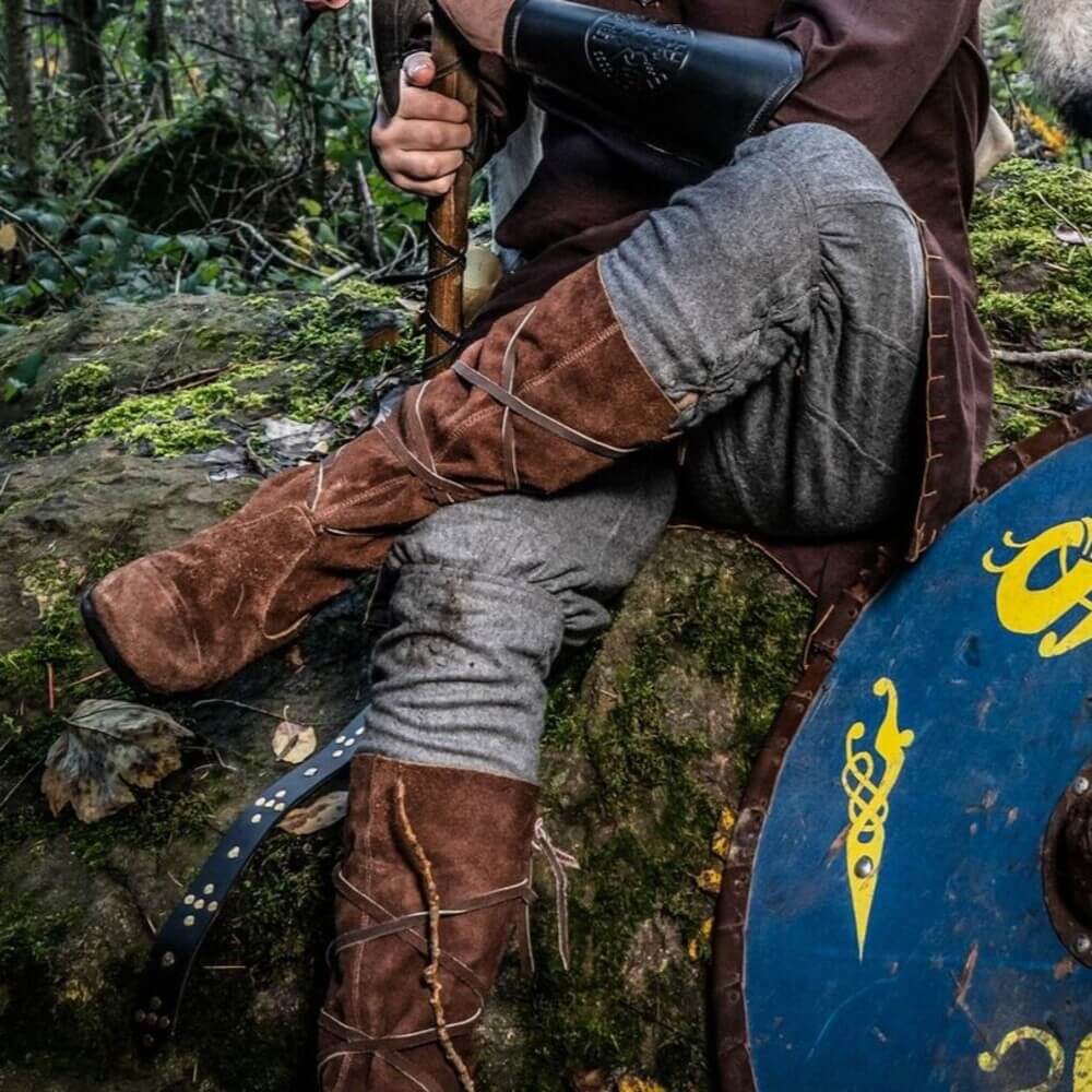 Person wearing brown suede renaissance wrap-tied boots, seated on mossy rock beside a blue Norse shield in a forest.