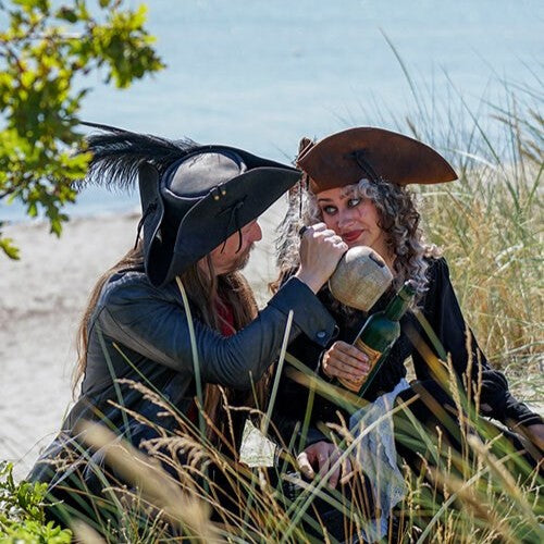 Two people in renaissance costumes by the beach; one wears a leather tricorn hat and holds a bottle.