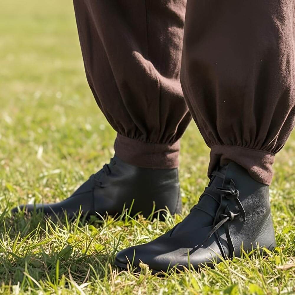 Black leather Renaissance ankle boots with four-string adjustment, worn with brown pants on grass.