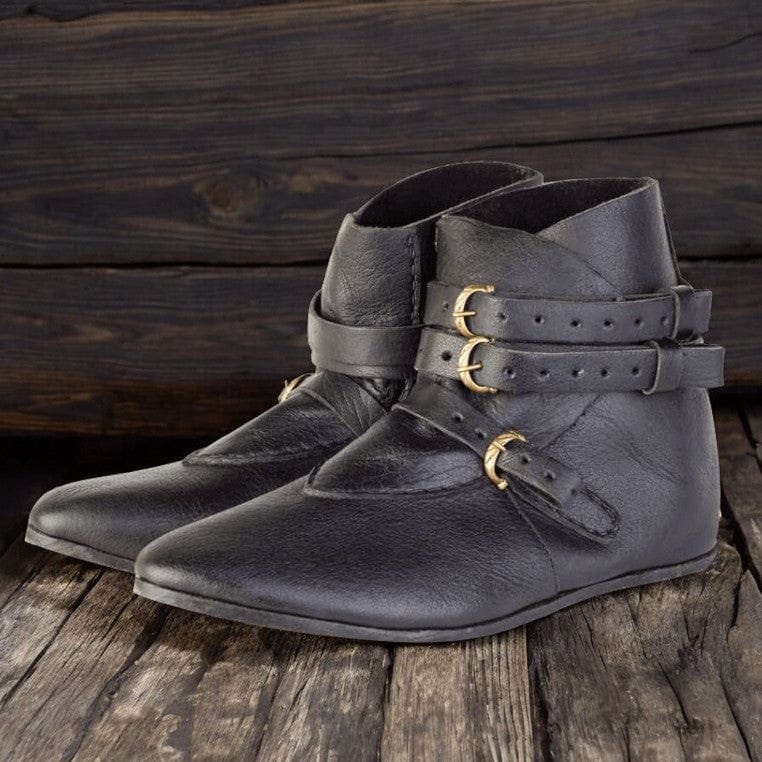 Black leather Renaissance ankle boots with three brass buckles on a dark wooden surface.