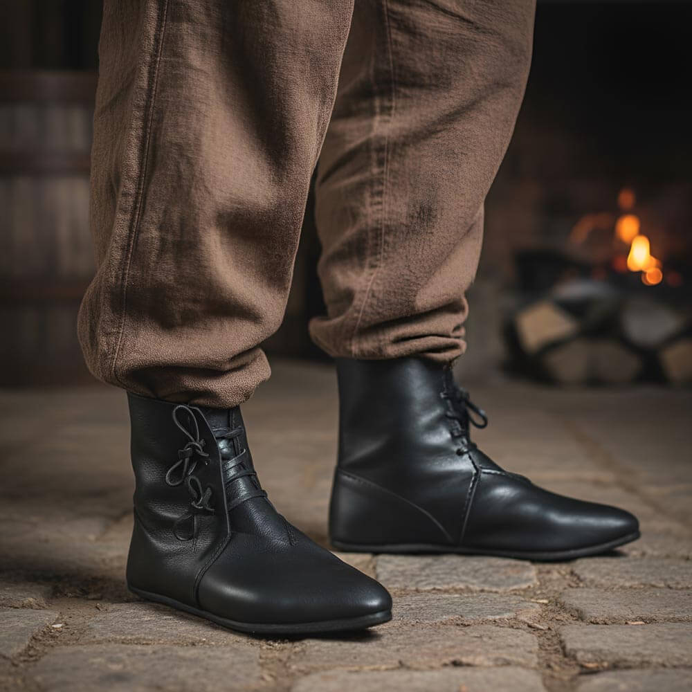 Person wearing Black Leather Renaissance Boots with Heinrich Ankle Design and brown pants standing in front of a fireplace.