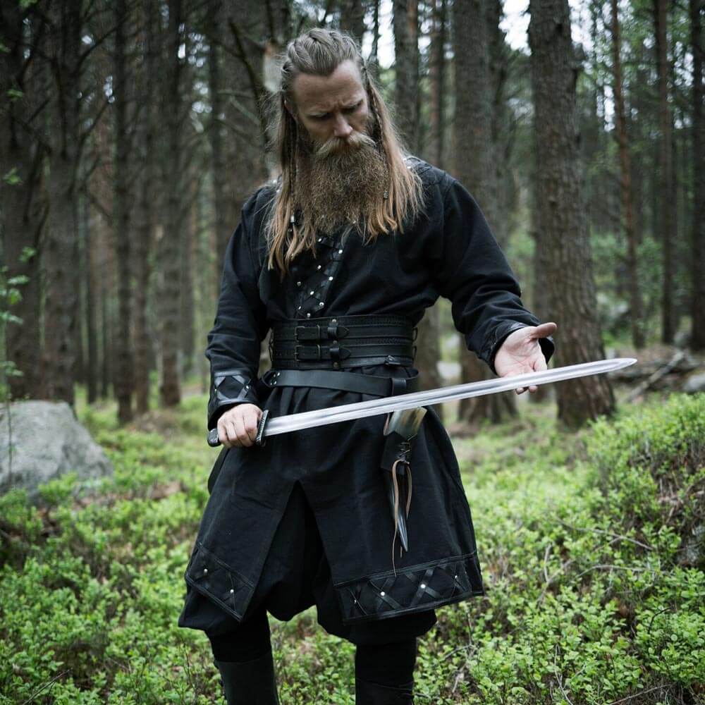 Man in a black Renaissance tunic with leather accents, holding a sword in a forest.