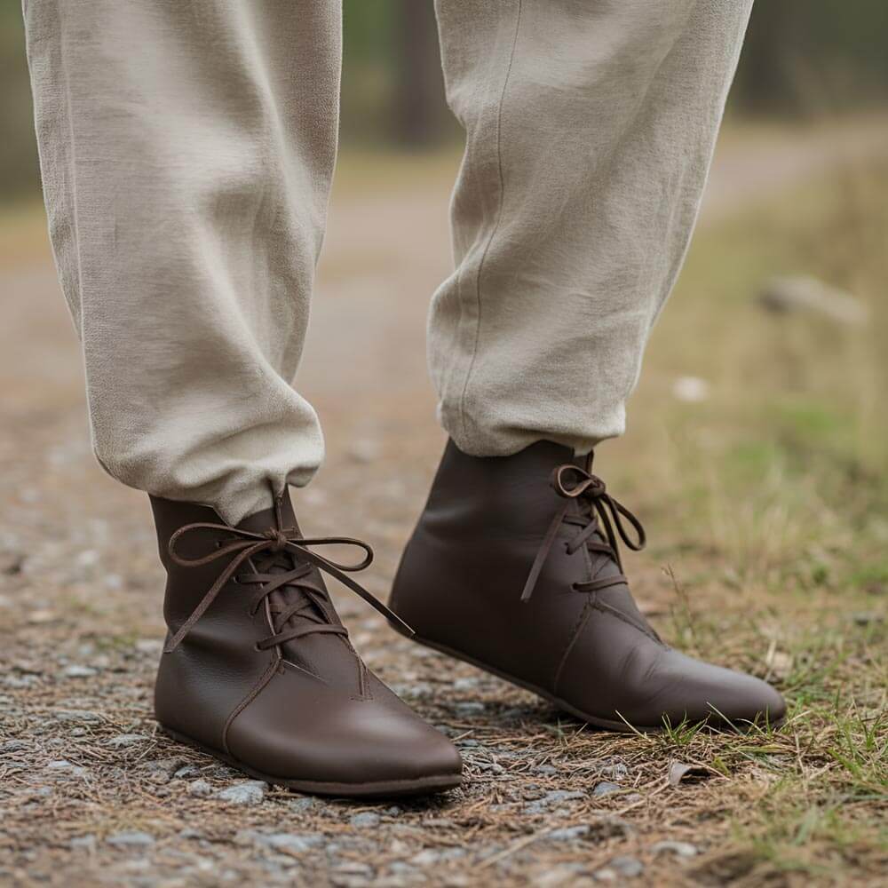 Brown leather Renaissance ankle boots with four-string adjustment and flat soles, worn on a dirt path.