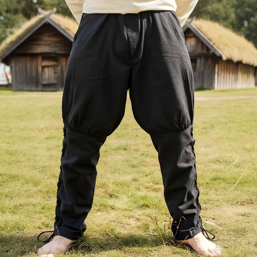 Person wearing black Renaissance pants standing on grass with wooden huts in the background