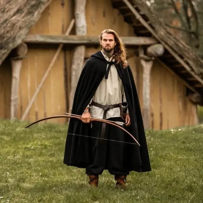 Man in a long black hooded Renaissance wool blend cloak, holding a bow in a grassy field.