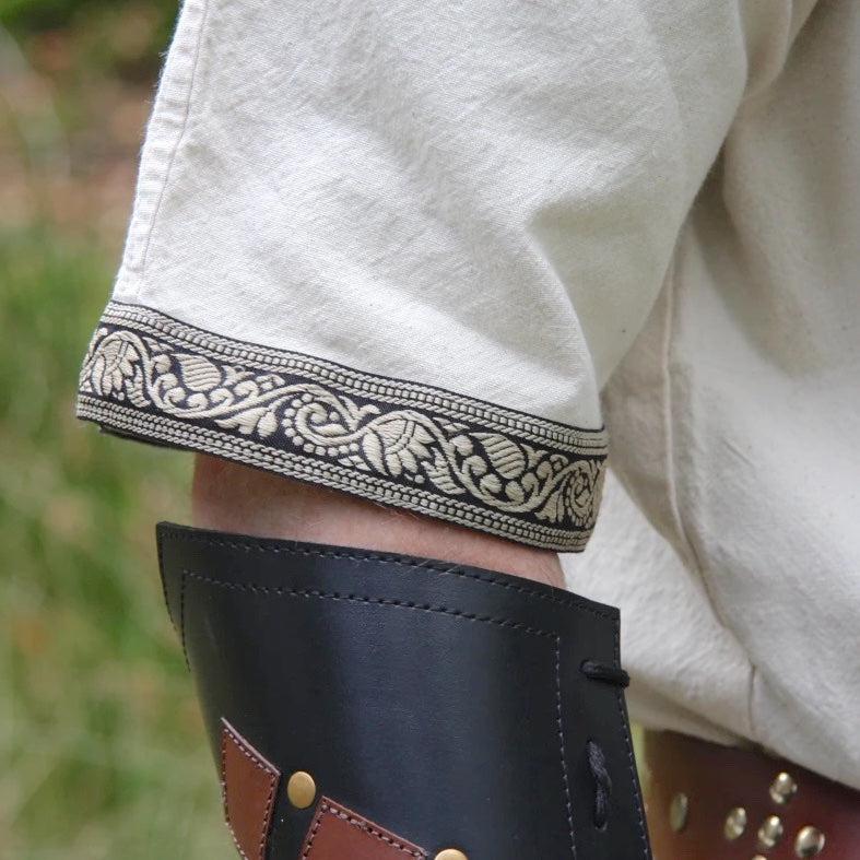 Natural off-white Renaissance cotton tunic sleeve with black and gold embroidered trim, worn with a leather bracer.