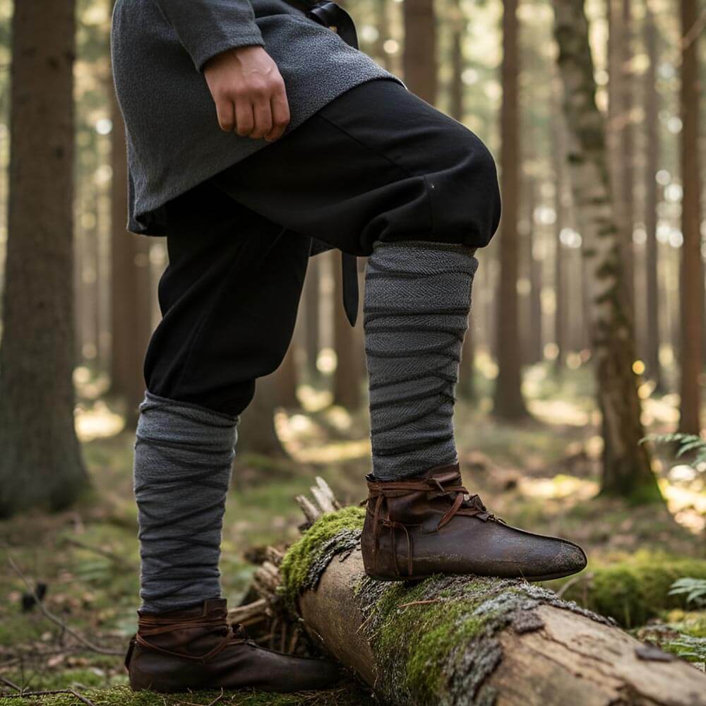 Person wearing grey Renaissance leg wraps, brown leather shoes and black pants, standing on a log in the forst.