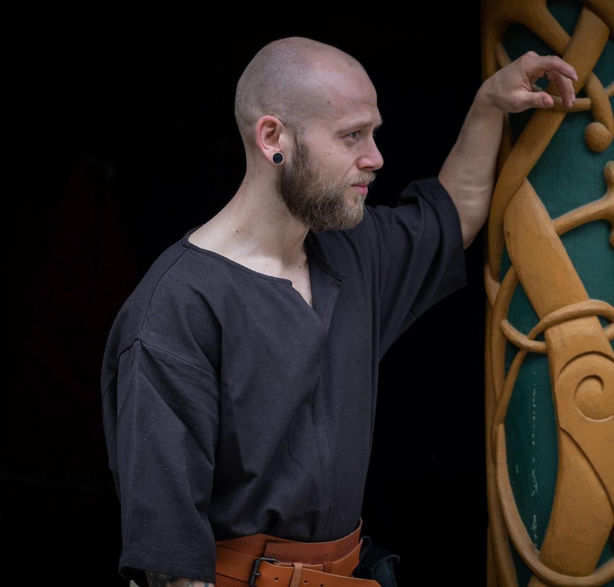 Renaissance short-sleeved black cotton tunic on a man, leaning against a carved wooden pillar.