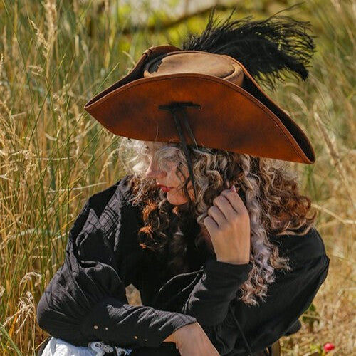 Brown leather Renaissance tricorn hat with black feather, worn by a person in tall grass.