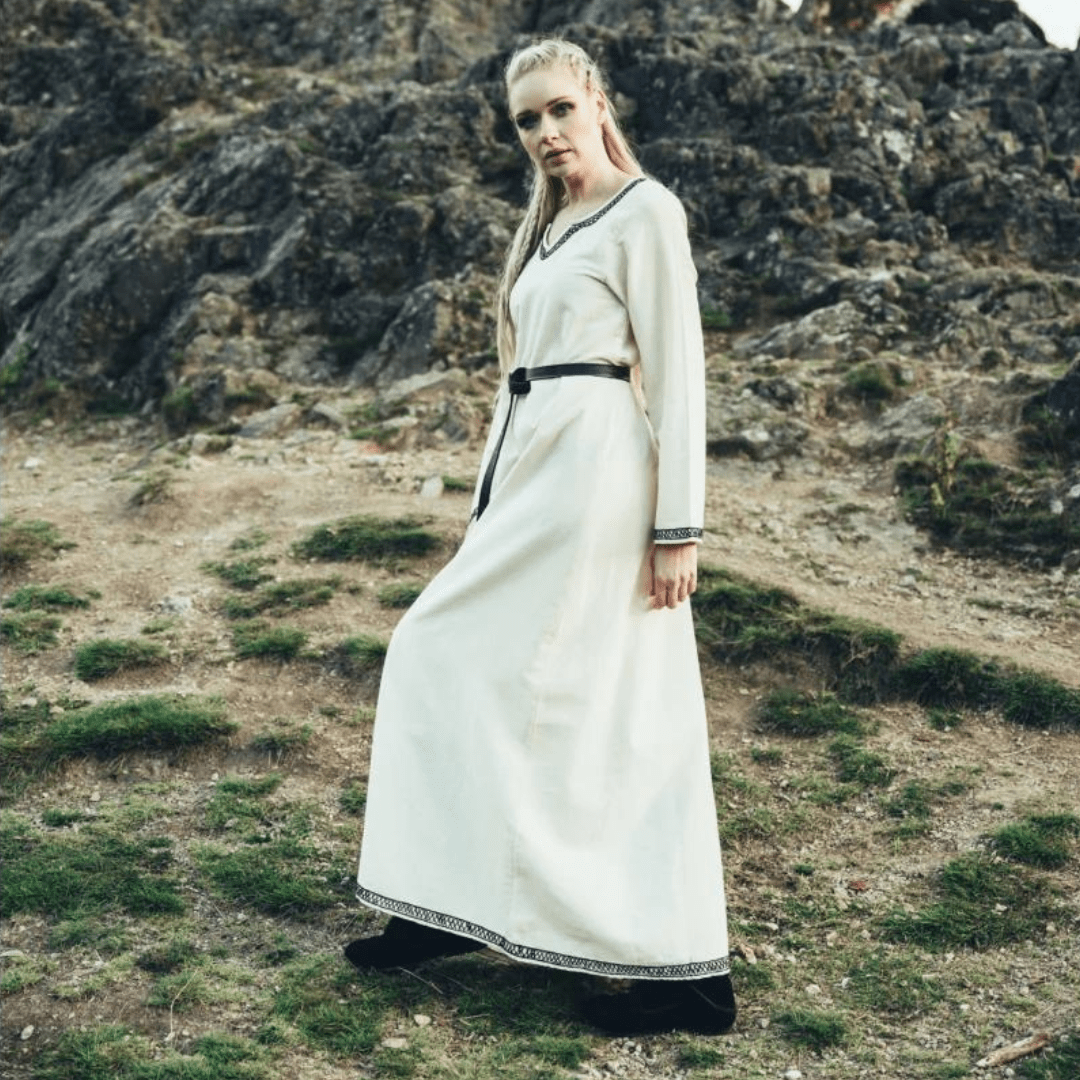Woman in a long, cream cotton Renaissance Viking dress with black embroidered trim and belt, standing outdoors.
